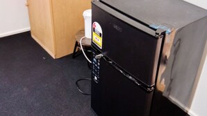 A compact black refrigerator with a control panel, a wooden cabinet with a white cup and a roll of paper towels, and a black carpeted floor. - Cozy sleep out  with free wi-fi (Kennington)