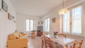 Un salon lumineux avec un canapé jaune, une table à manger recouverte d’une nappe à motifs floraux et un téléviseur en arrière-plan.