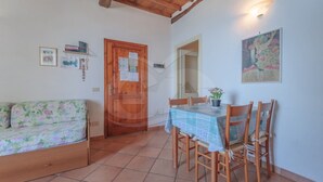 Un comedor con una mesa dispuesta para cuatro personas, rodeada de sillas de madera. Un sofá floral se encuentra a la izquierda, y se ve una puerta de madera con un tablón de anuncios.
