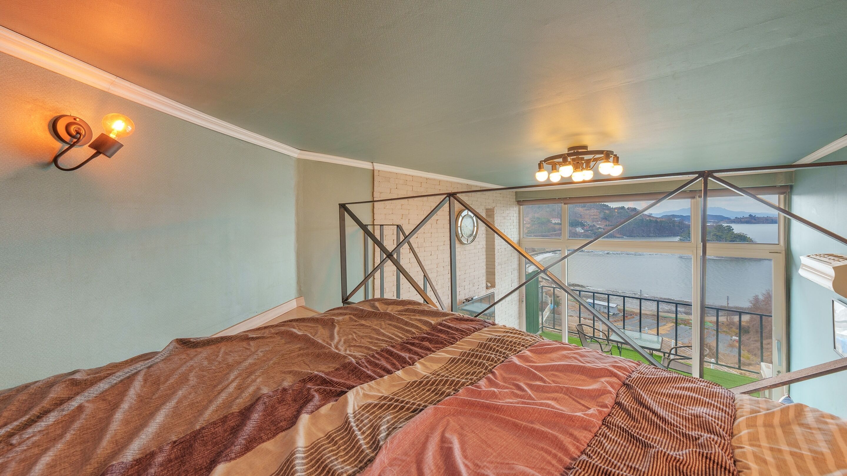 A bedroom with a bed, a window with a view of a body of water, and a balcony railing.