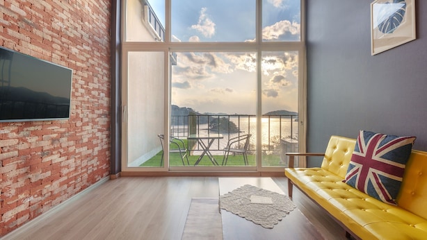 A modern living room with a brick wall, a yellow sofa with a Union Jack pillow, a large window with a view of a body of water and hills, and a flat-screen TV mounted on the wall. - Yeosu Island Pet Pension (Yeosu)