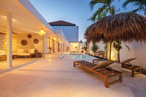 Un área de piscina moderna al aire libre con sillones de descanso, comedor y una estructura de techo de paja.