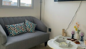 A living room with a grey sofa, a small round table with yellow legs, a floor lamp, and a flat-screen TV mounted on the wall.