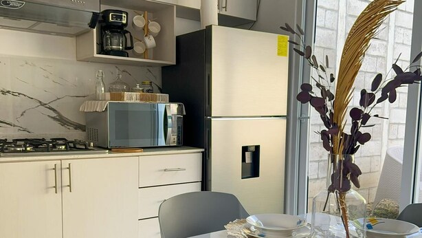 A modern kitchen with white cabinets, a built-in microwave, and a refrigerator. There is a dining table with plates and a vase of dried plants.