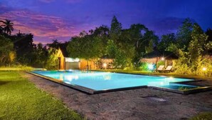 A swimming pool with a stone border, surrounded by grass and trees, and a thatched-roof structure in the background. - Happy Man Village (Kosgoda)