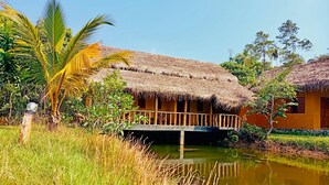 A thatched-roof house with a wooden deck over a pond, surrounded by tropical vegetation. - Happy Man Village (Kosgoda)