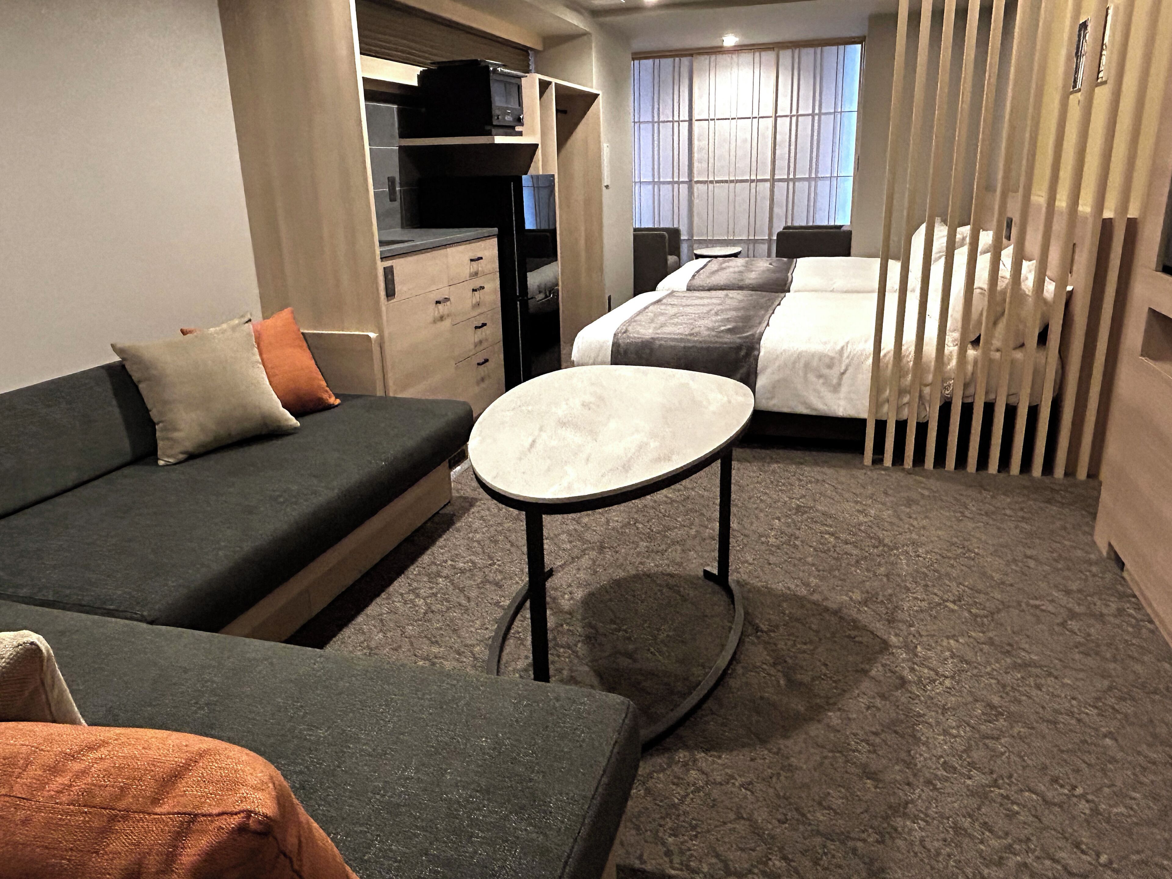 A compact hotel room with a sofa, a small round table, a bed with a white duvet, and a window with vertical blinds.