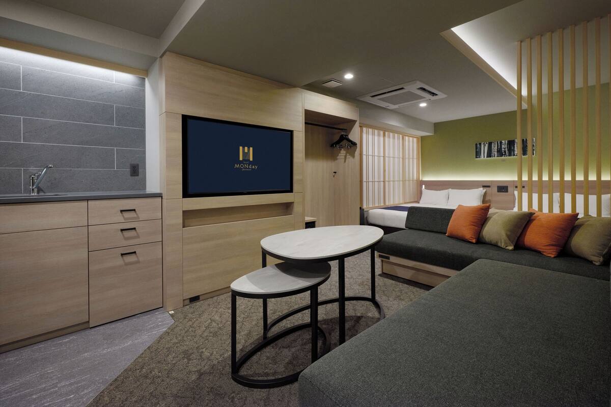 Habitación de hotel moderna con kitchenette, televisor de pantalla plana montado en la pared, sofá con cojines y una pequeña mesa redonda.