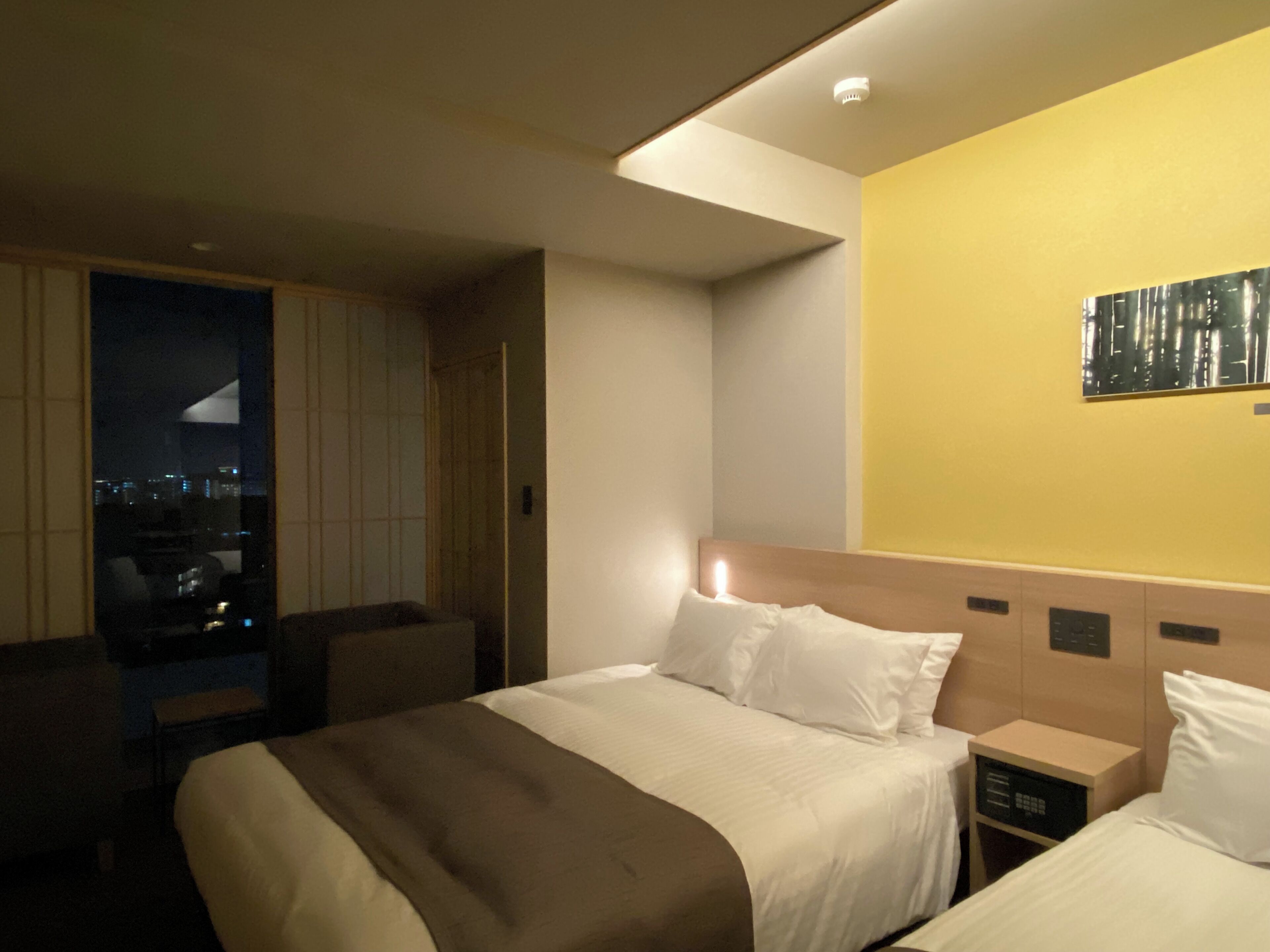 A hotel room with a bed, bedside tables, and a cityscape view through a window.