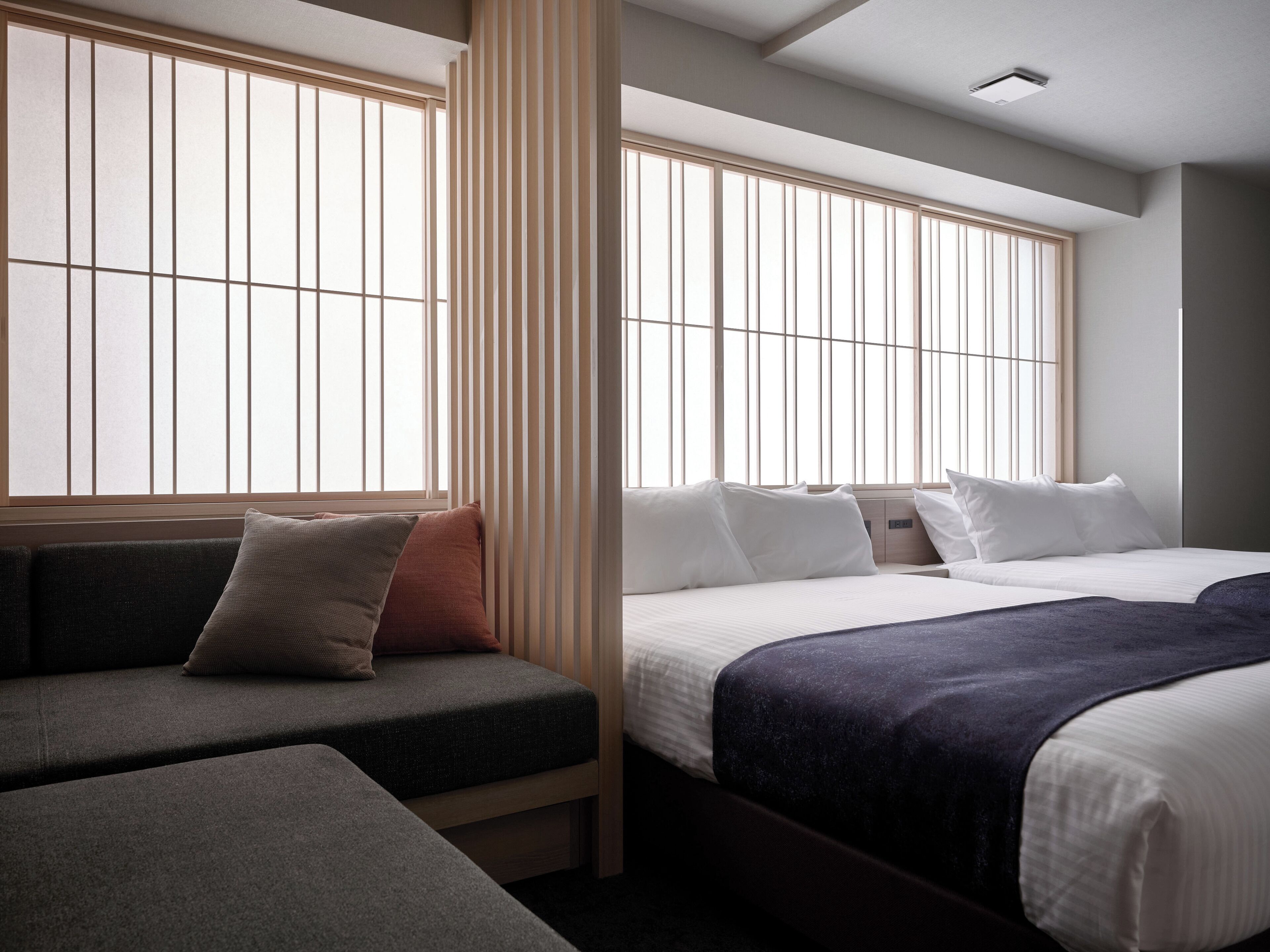 A hotel room with a sofa, two beds, and large windows with vertical blinds.
