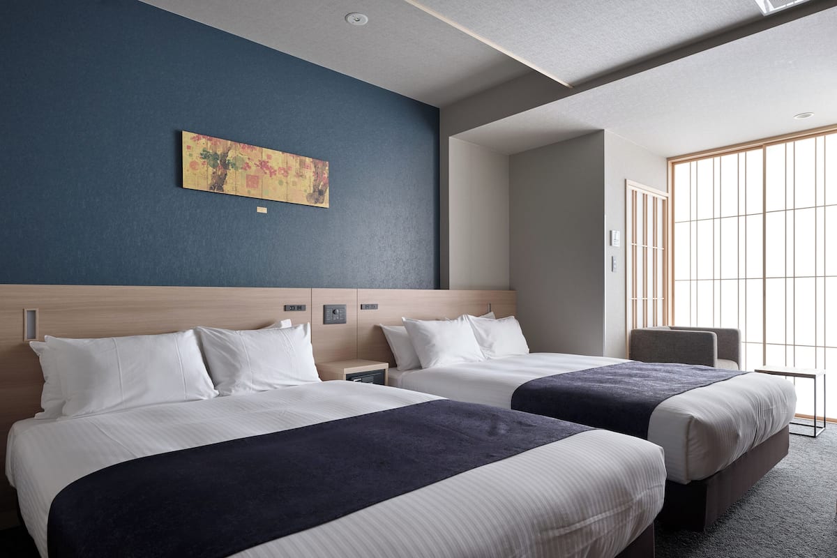 Habitación de hotel con dos camas, cabeceros, mesitas de noche y una silla. Cuenta con una ventana con persianas y una obra de arte colgada en la pared.