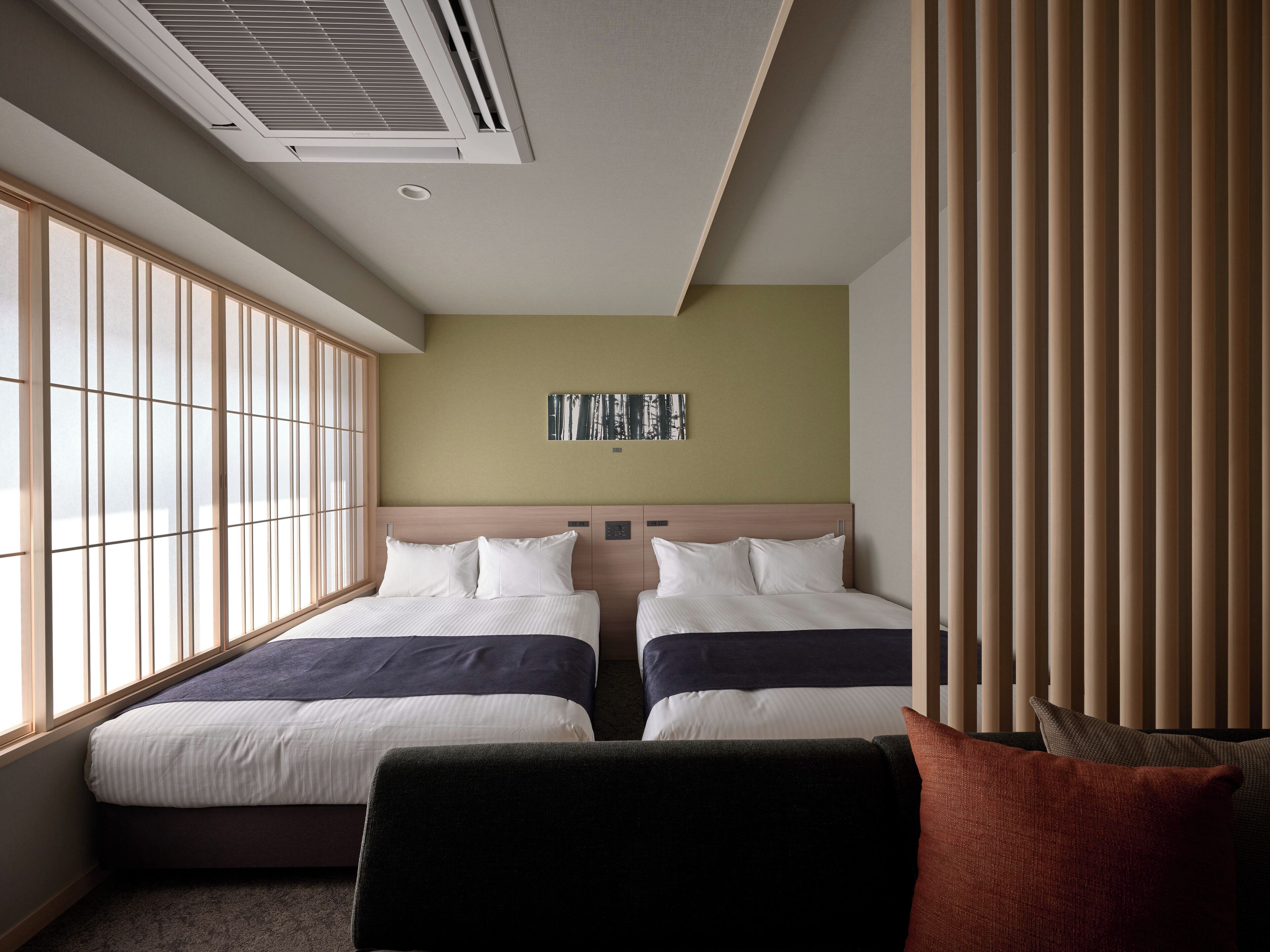 A hotel room with two beds, a large window, a ceiling vent, and a wall-mounted artwork.