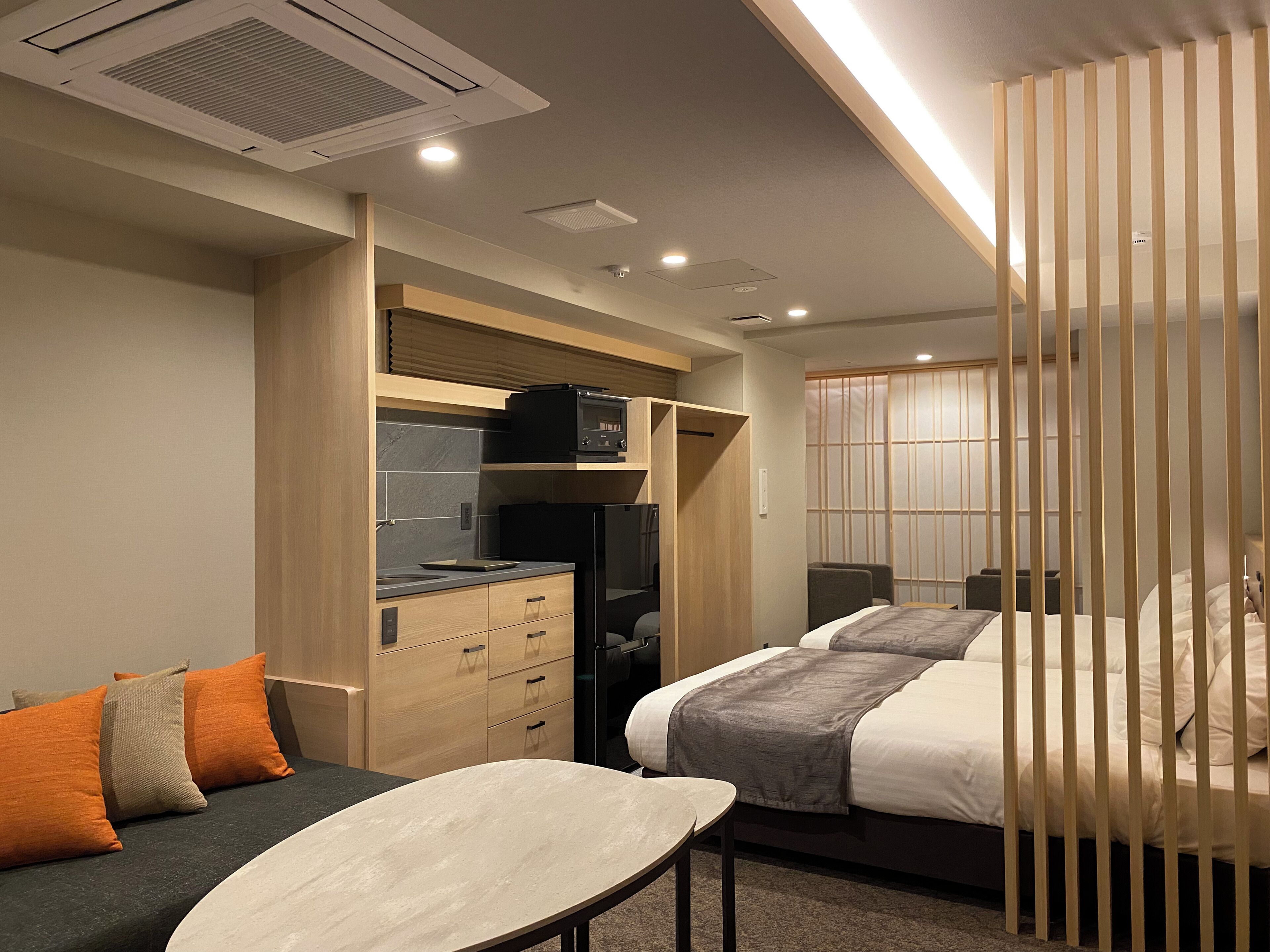 A modern hotel room with a kitchenette, two beds, a desk, and a sliding door.