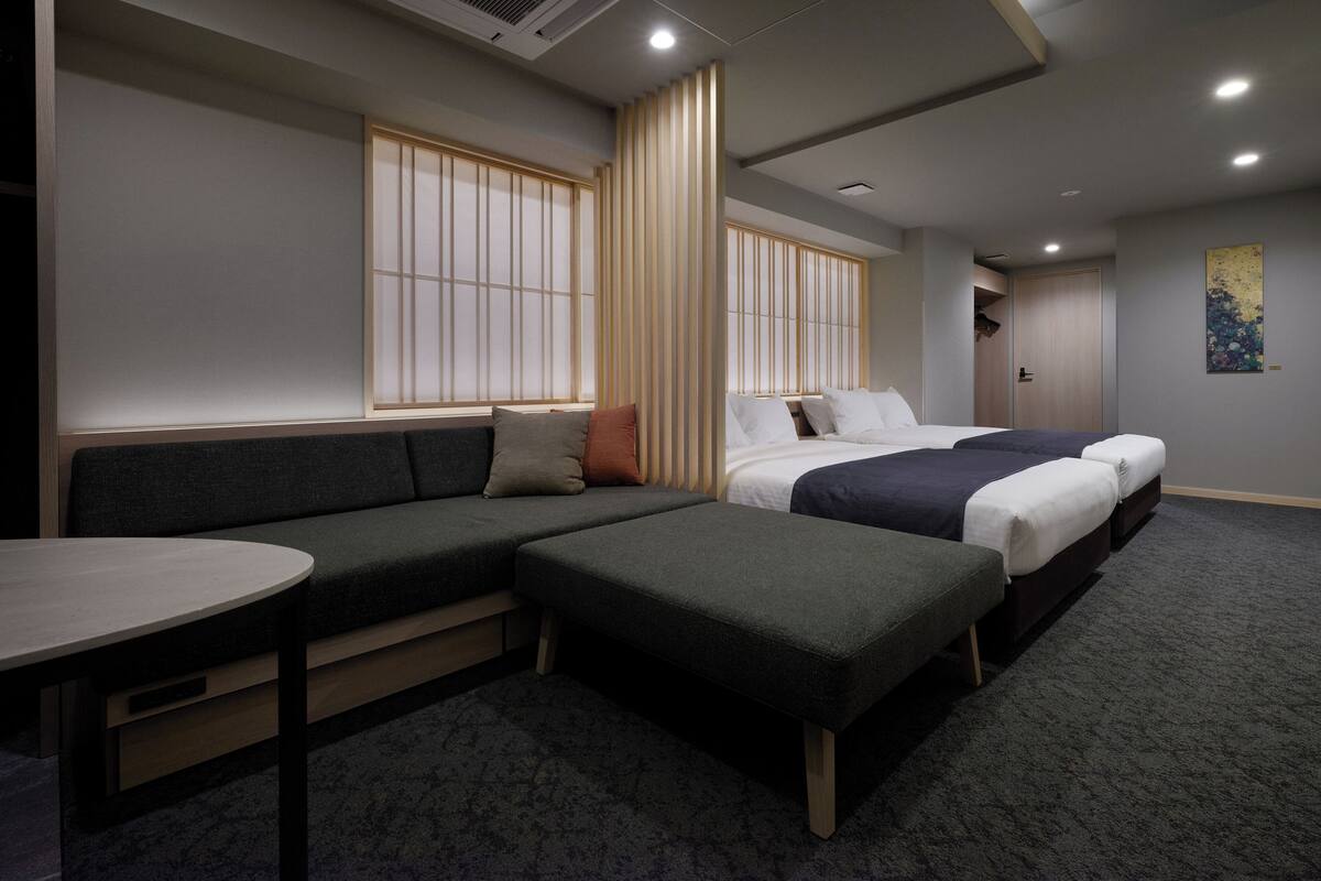 Una habitación de hotel moderna con sofá, dos camas individuales, una mesita pequeña y un amplio ventanal con persianas verticales.