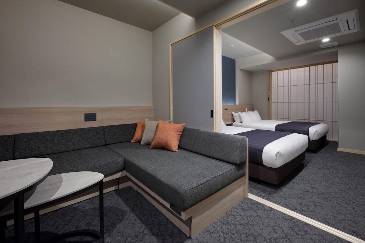 Una habitación de hotel moderna con un sofá seccional gris, una pequeña mesa redonda, dos camas individuales y un espejo grande que refleja la habitación.