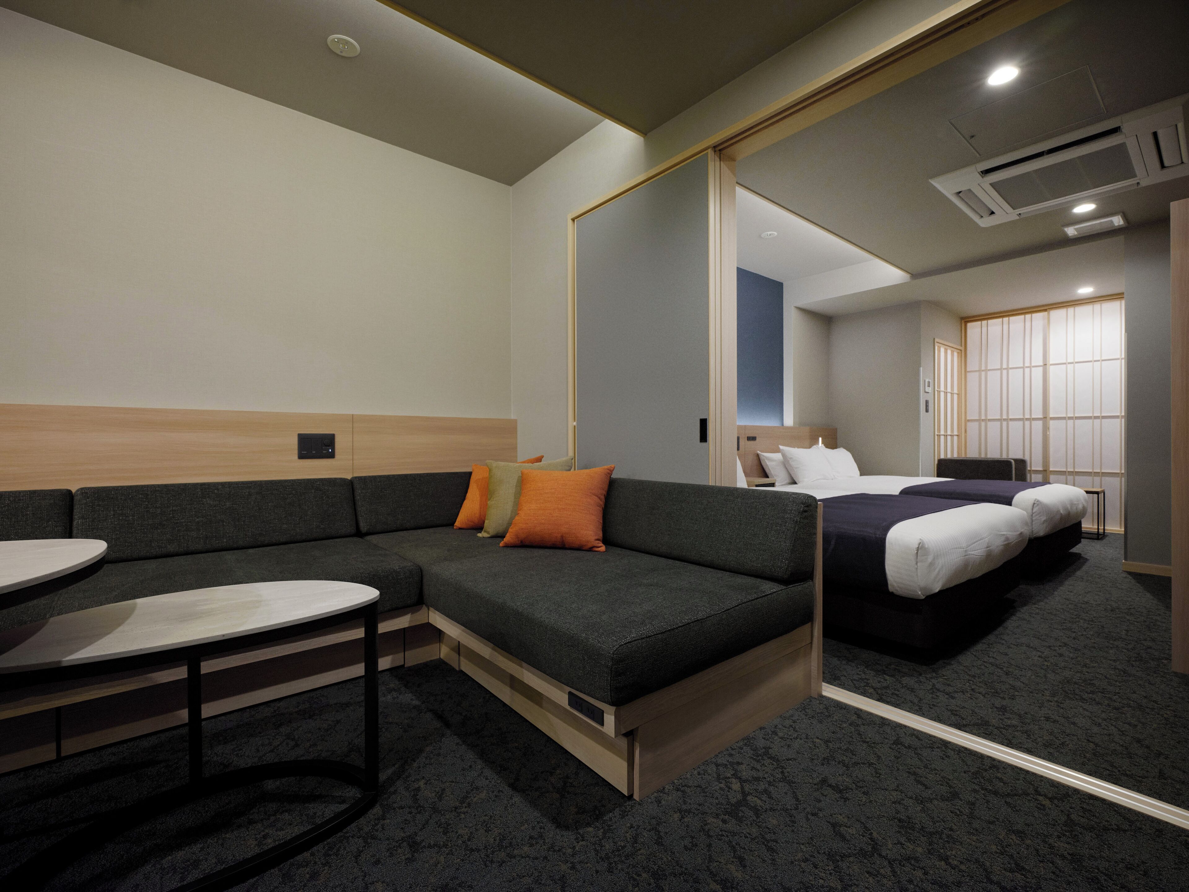 A modern hotel room with a sectional sofa, a small round table, and a bed with two pillows.