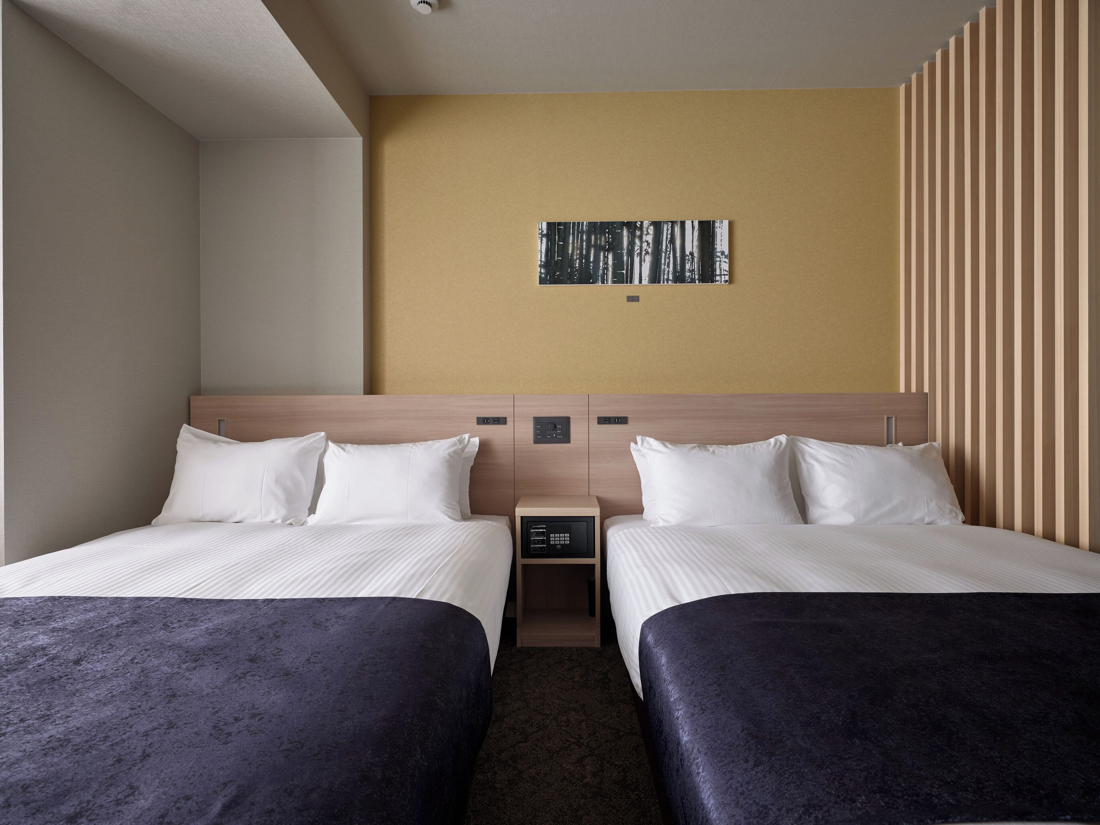 A hotel room with two beds, a small bedside table, a wall-mounted control panel, and a framed picture on the wall.