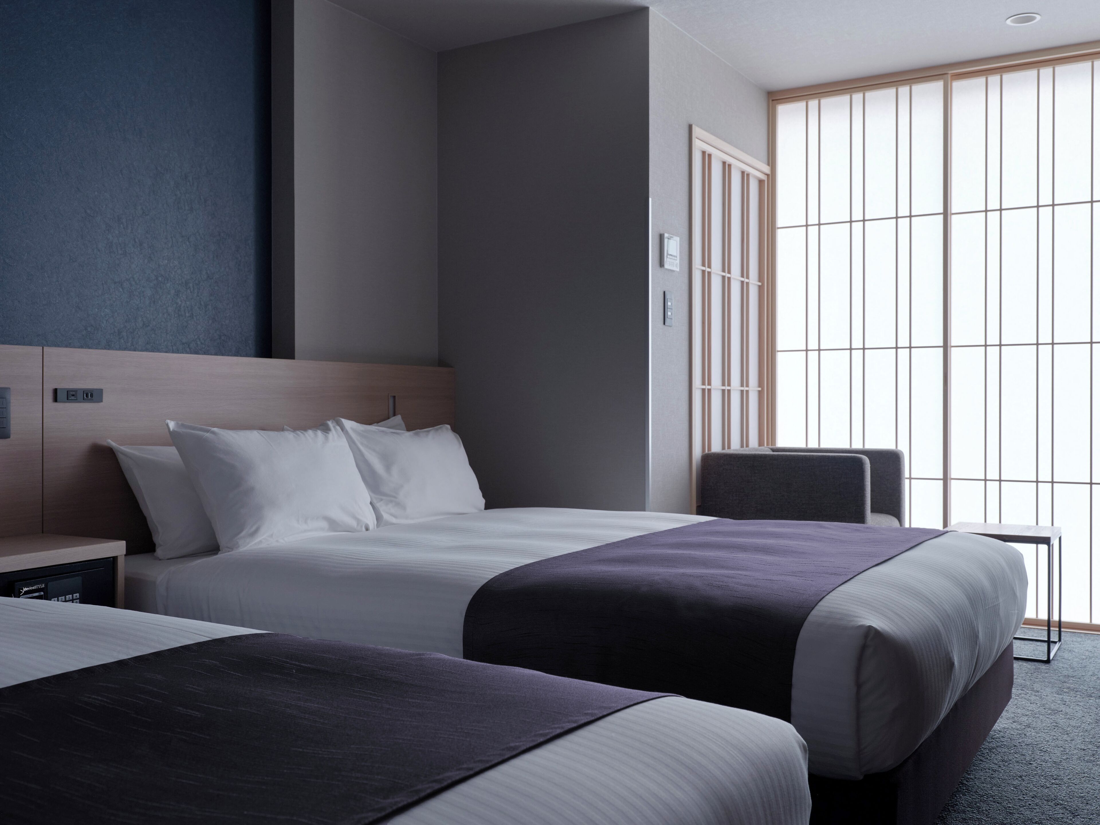 A hotel room with two beds, a wooden headboard, a nightstand, a desk, and a large window with vertical blinds.