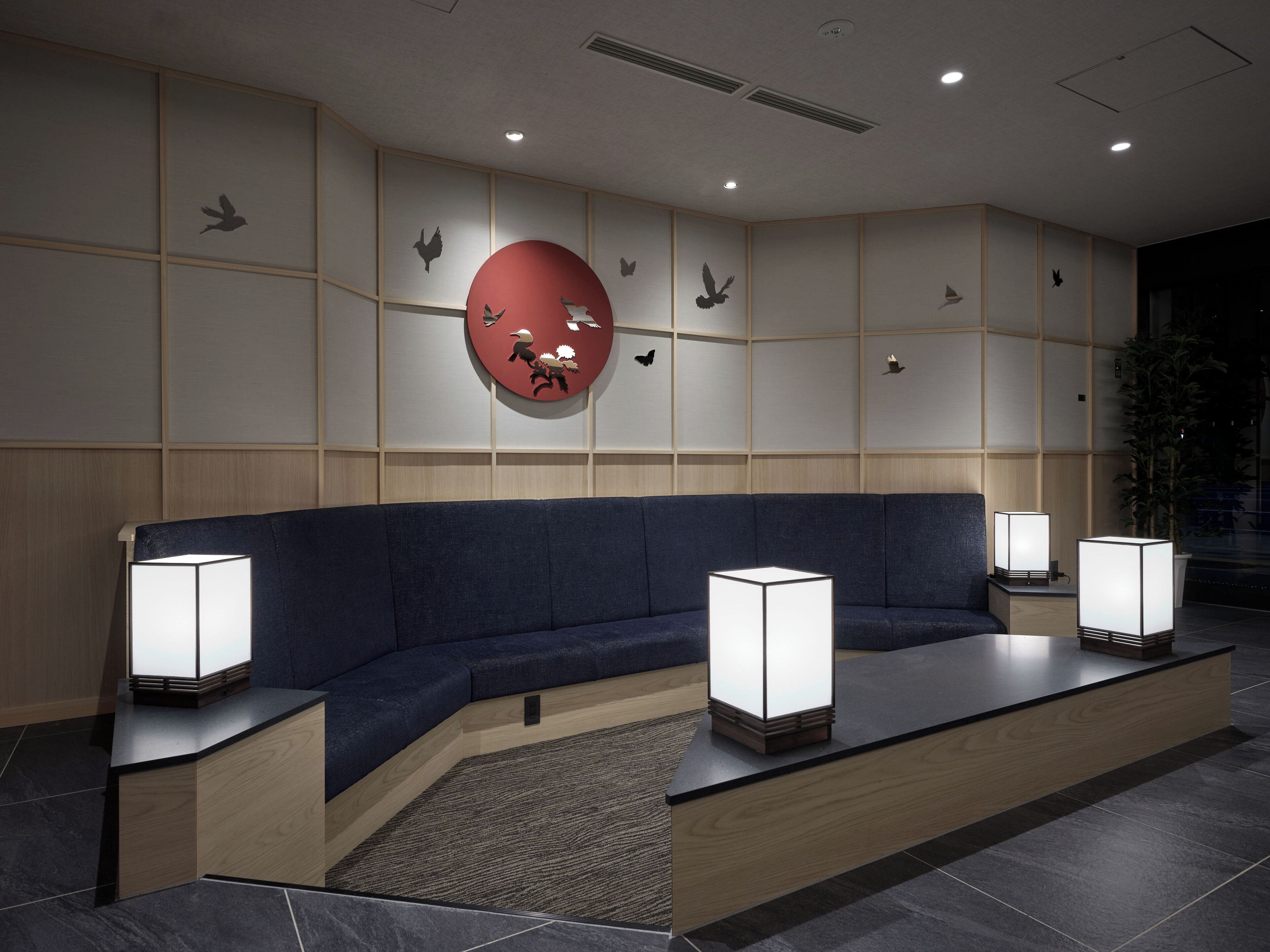Un espace d’attente moderne avec un canapé bleu foncé, deux lampadaires et une grande décoration murale circulaire rouge.