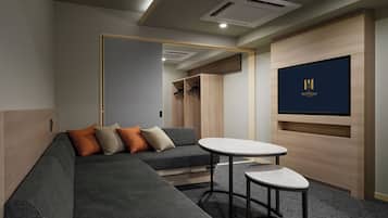 A modern hotel room with a grey sectional sofa, a small round table, and a flat-screen TV mounted on the wall.