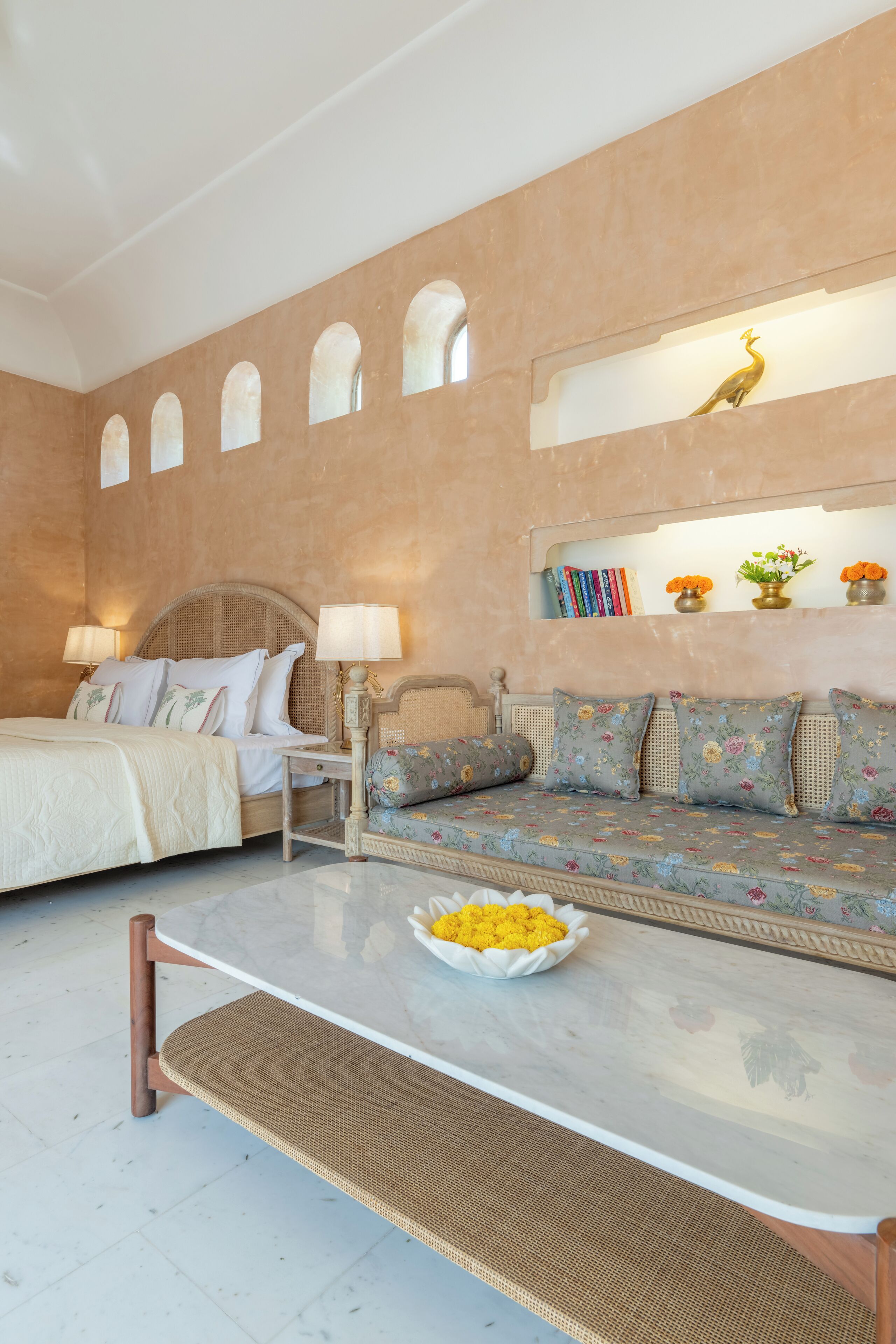 A bedroom with a bed, a sofa, a table with a bowl of fruit, and a bookshelf with books and flowers.