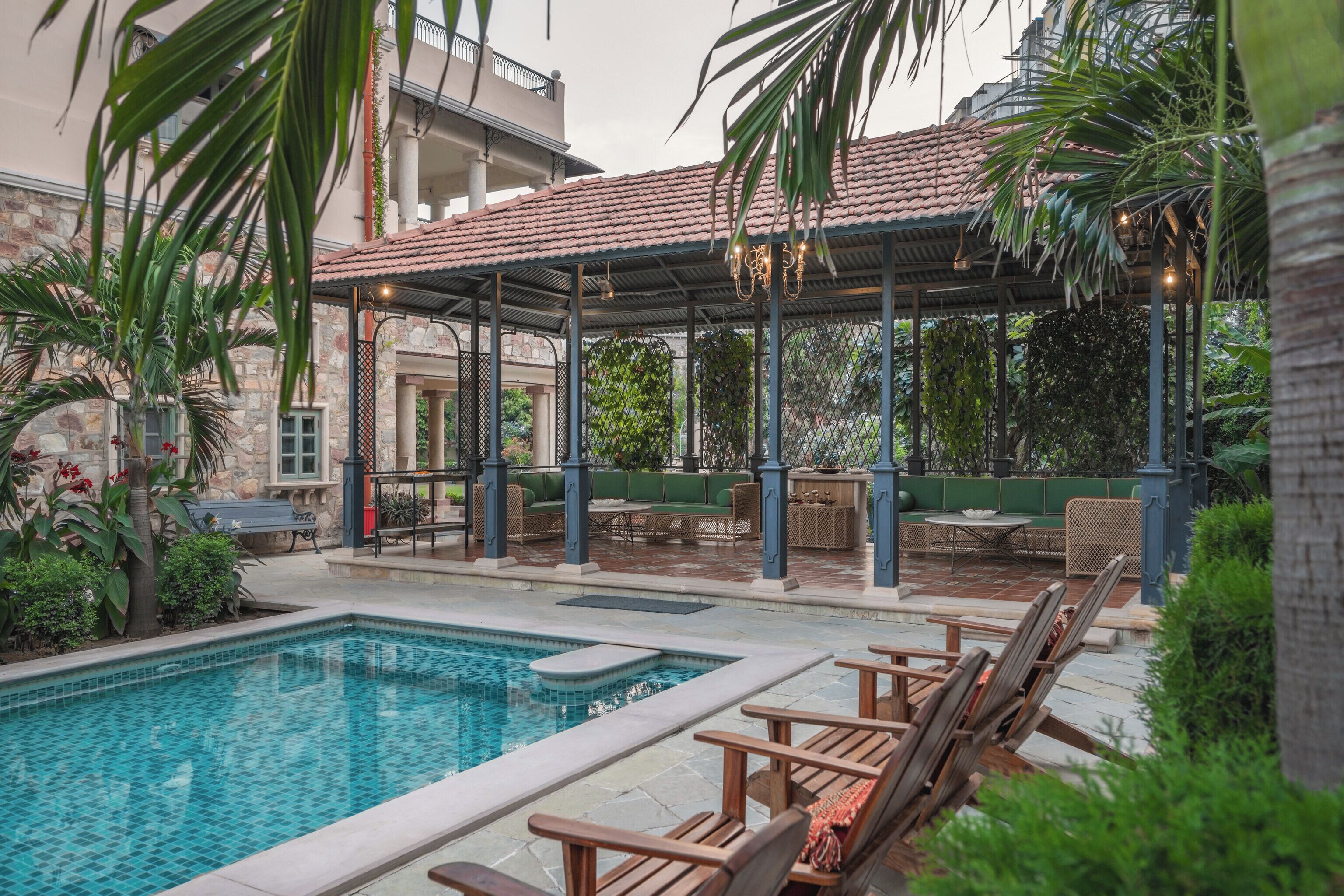 Zona de piscina con área de descanso cubierta, sillas reclinables y un edificio de piedra al fondo.