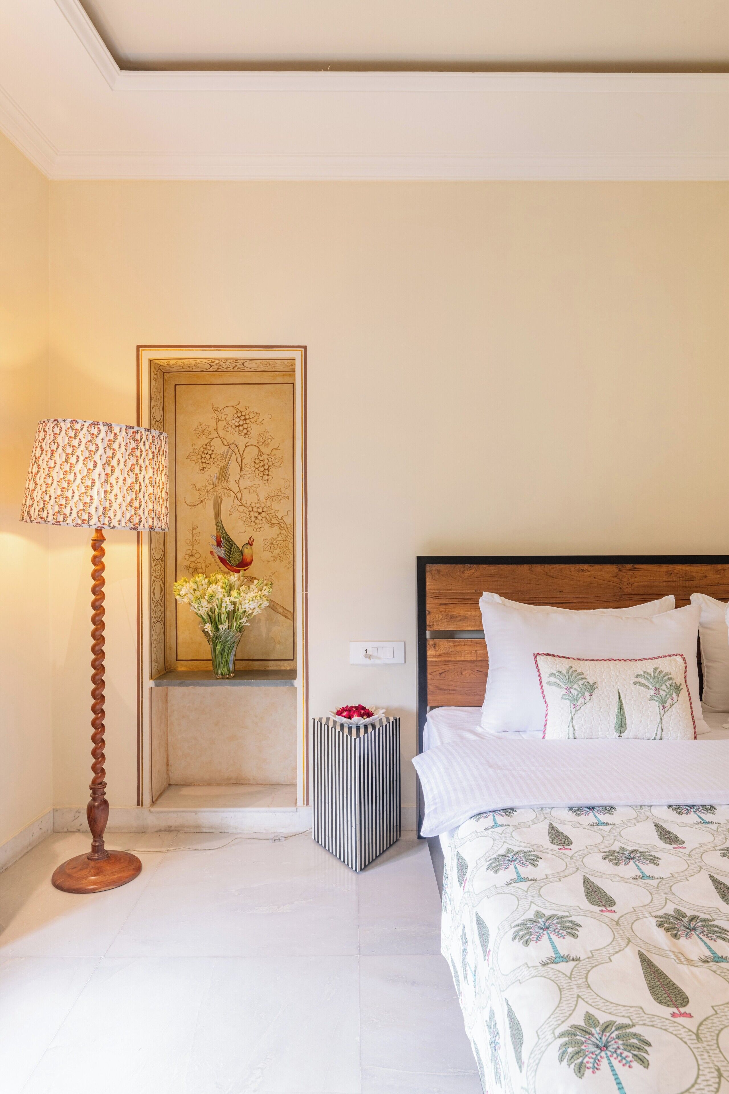 A hotel room with a bed, a lamp, a decorative wall piece, and a small table with a vase of flowers.