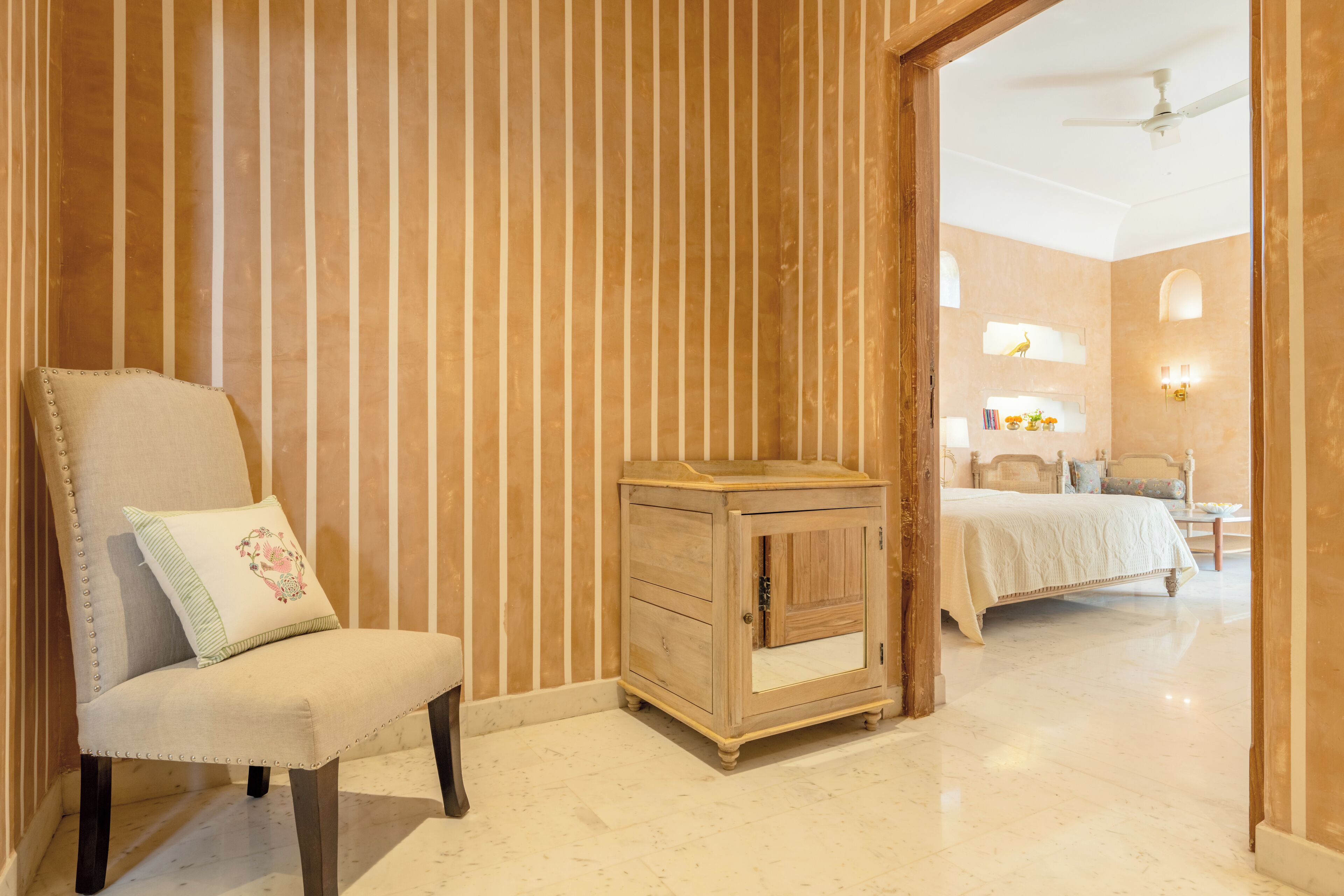 A room with a striped wall, a wooden cabinet, a bed, and a chair with a pillow.