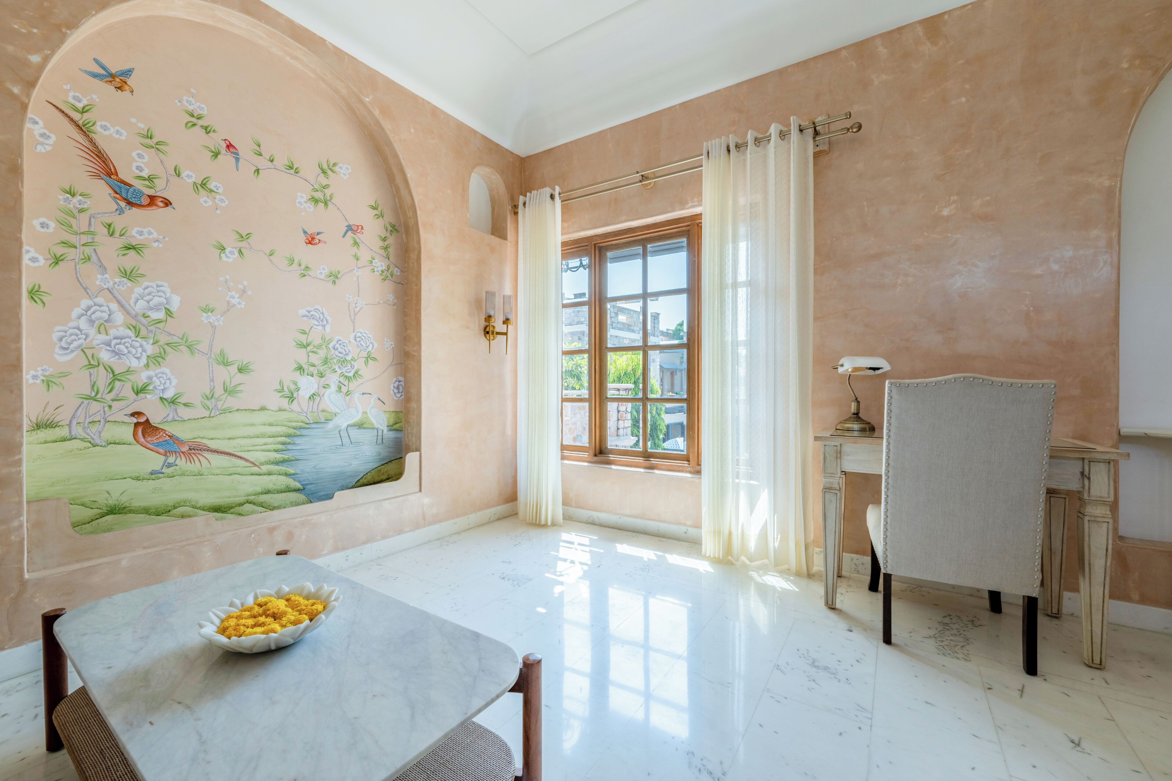 A room with a mural of birds and flowers, a dining table with a bowl of fruit, a window with curtains, and a chair.