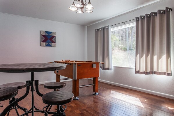 Una habitación con una mesa de madera y taburetes, una mesa de futbolín, un cuadro en la pared y una ventana con cortinas.