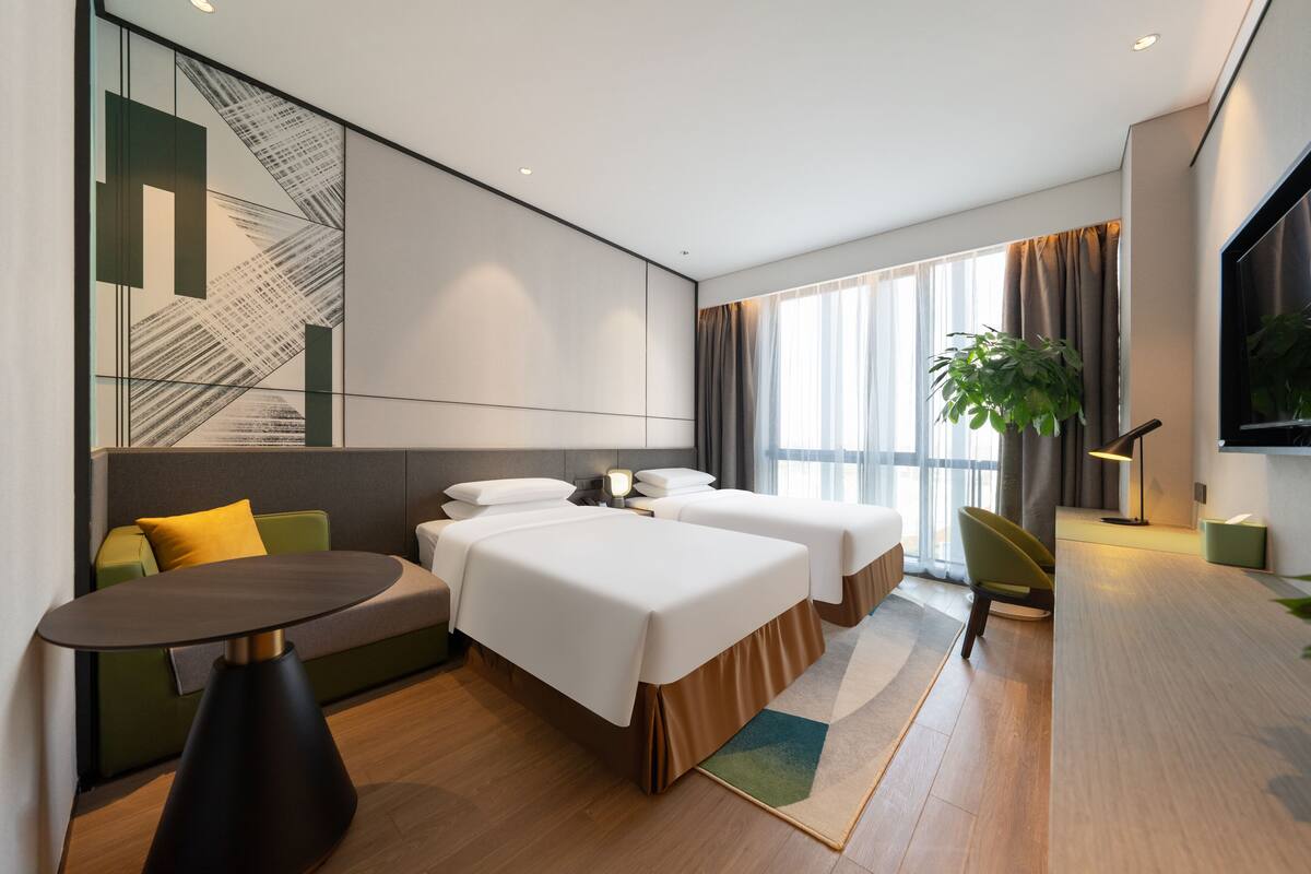 A modern hotel room with two beds, a sofa, a round table, and a large window with curtains.