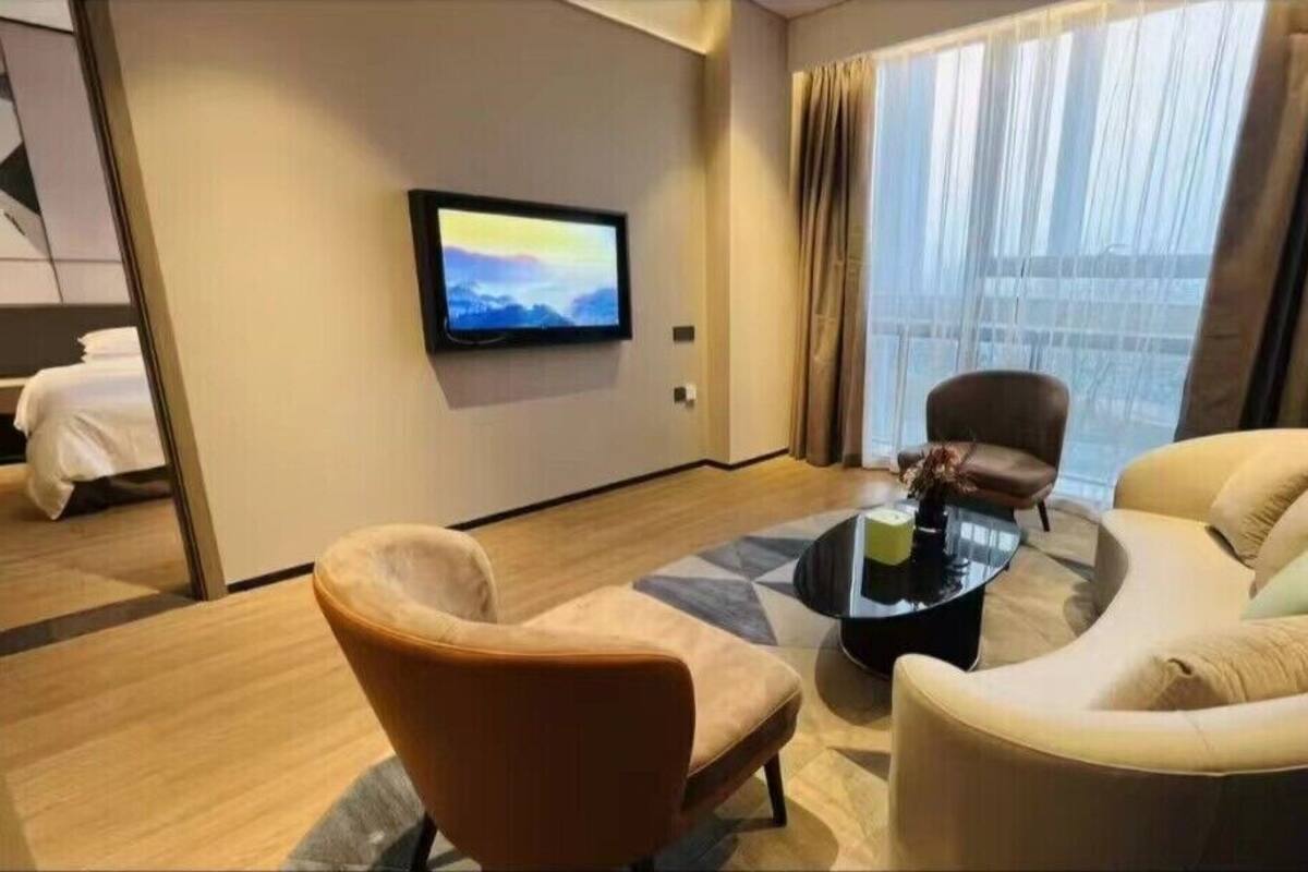 A modern hotel room with a sofa, armchair, and a flat-screen TV mounted on the wall.