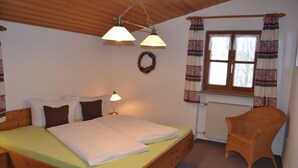 Ein Schlafzimmer mit einem hölzernen Bett, zwei Nachttischlampen, einem Korbstuhl, einem Fenster mit Vorhängen und einer Wandkrone.