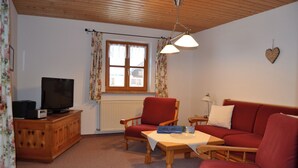 A living room with a television, wooden furniture, a window with curtains, and a heart-shaped decoration on the wall. - Apartment 2 - Göllblick, for 1-4 persons, approx. 50 sqm, 1 bedroom, living room/bedroom, kitchen, shower, WC, balcony (Bischofswiesen)