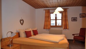 A bedroom with a wooden bed, a desk with a lamp, a chair, and a window with curtains. - Apartment 2 - Göllblick, for 1-4 persons, approx. 50 sqm, 1 bedroom, living room/bedroom, kitchen, shower, WC, balcony (Bischofswiesen)