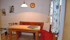 A small, cozy room with a wooden table and bench, a clock on the wall, and a window with floral curtains. - Apartment 2 - Göllblick, for 1-4 persons, approx. 50 sqm, 1 bedroom, living room/bedroom, kitchen, shower, WC, balcony (Bischofswiesen)