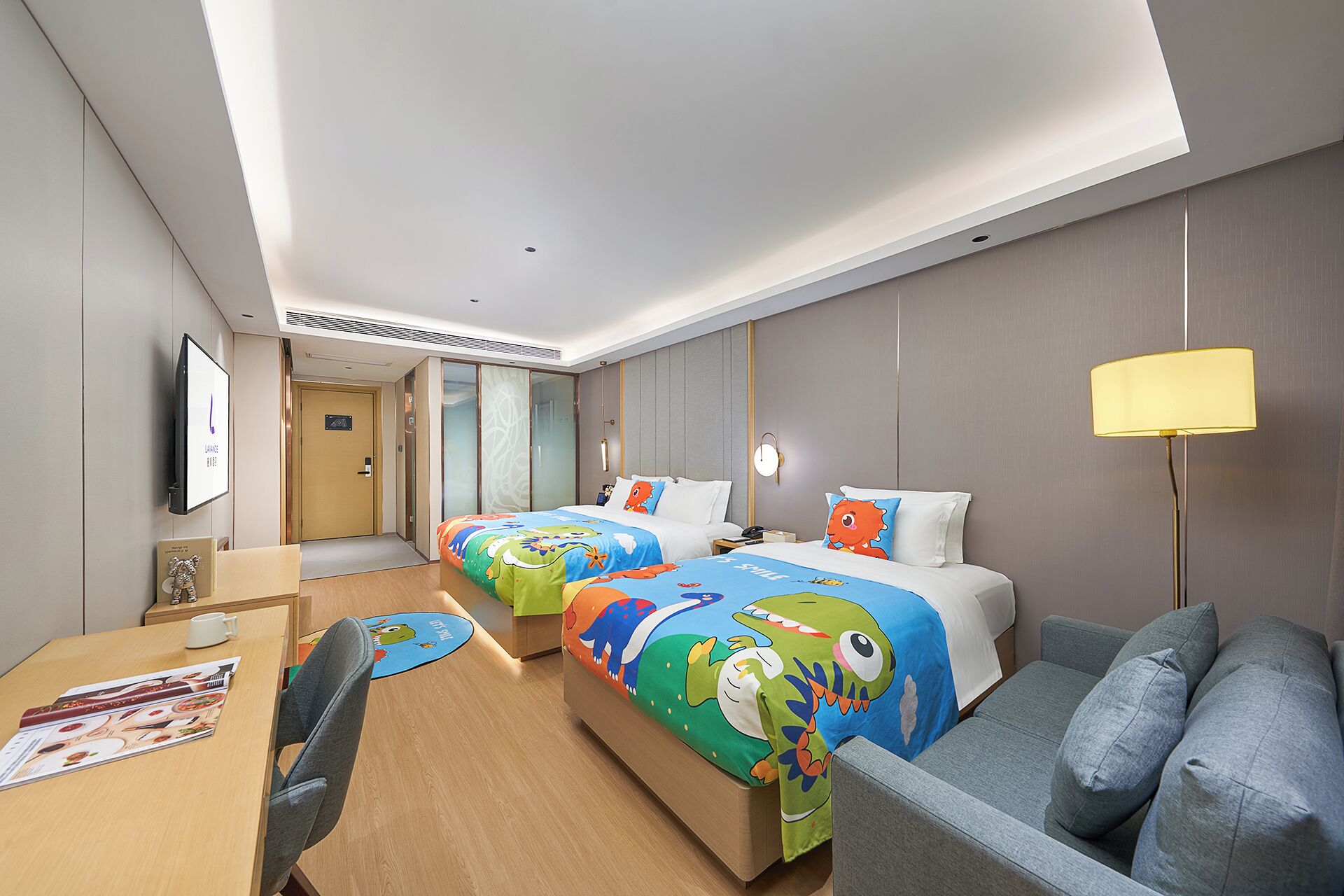A hotel room with two beds, a desk, a chair, a sofa, a TV, and a lamp.