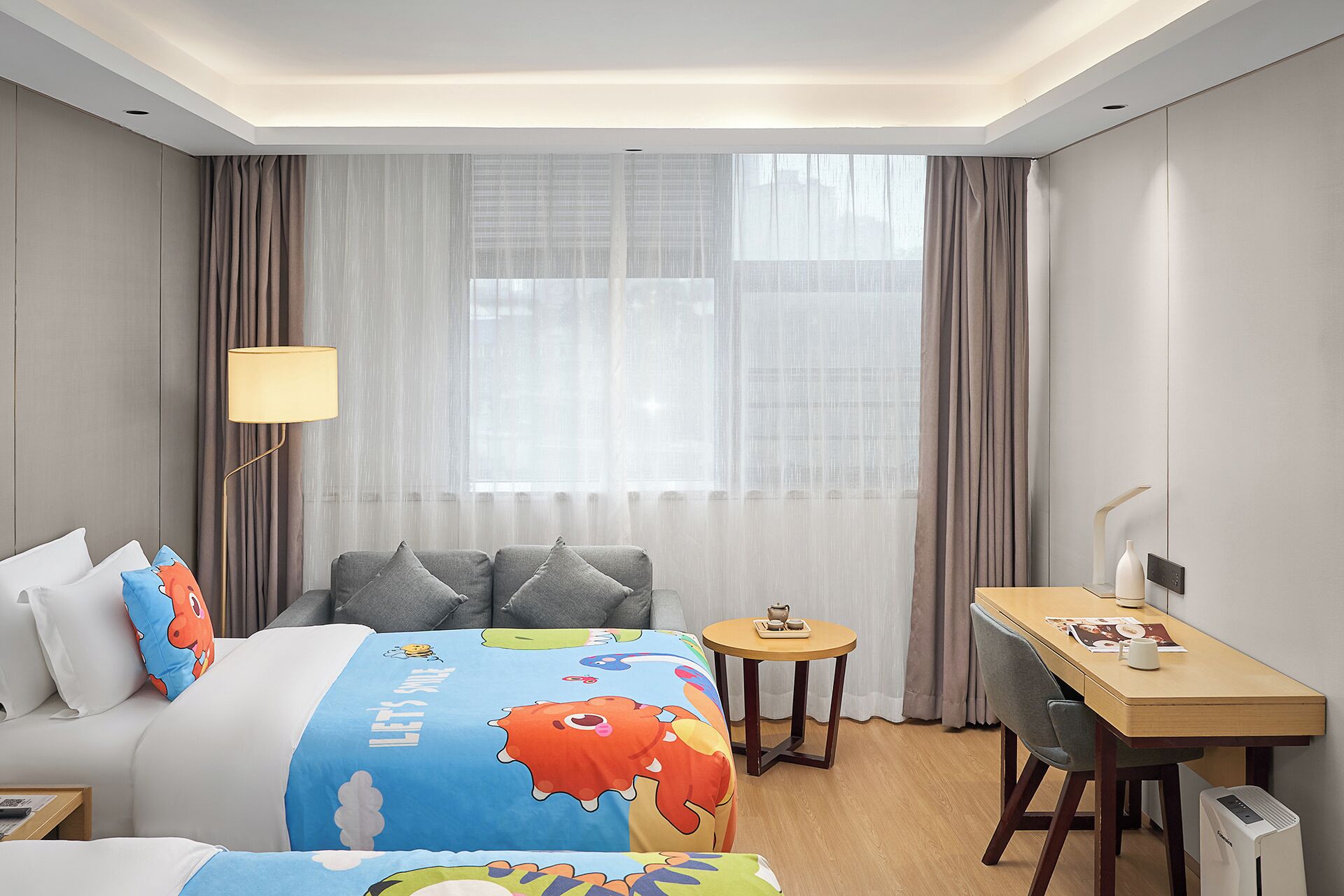 A hotel room with a bed, a desk, a lamp, and a view of the outside through sheer curtains.