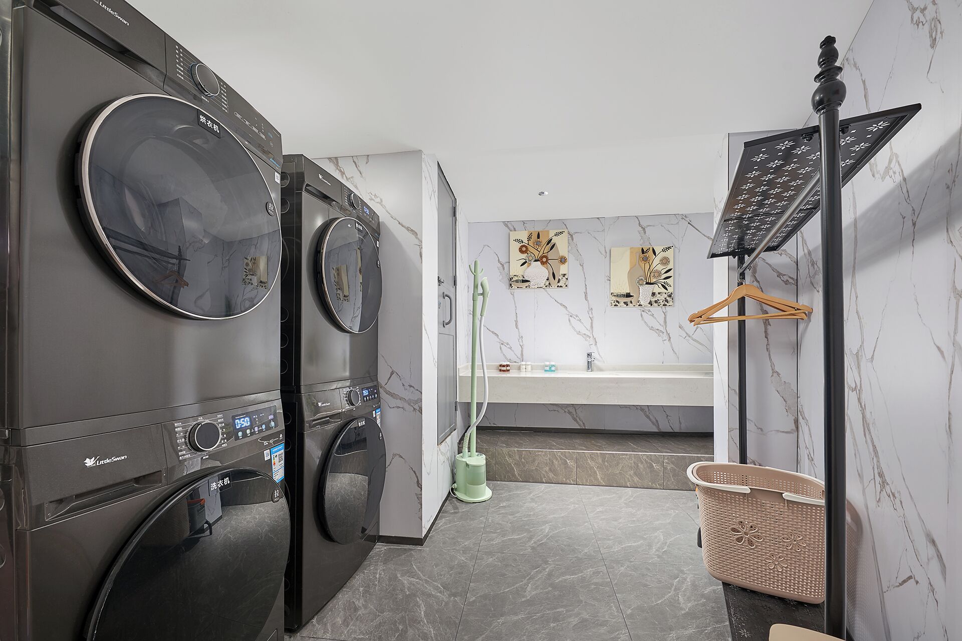 A modern laundry room with two stacked front-loading washing machines, a drying rack, a hanging clothes rack, and a marble-patterned wall.