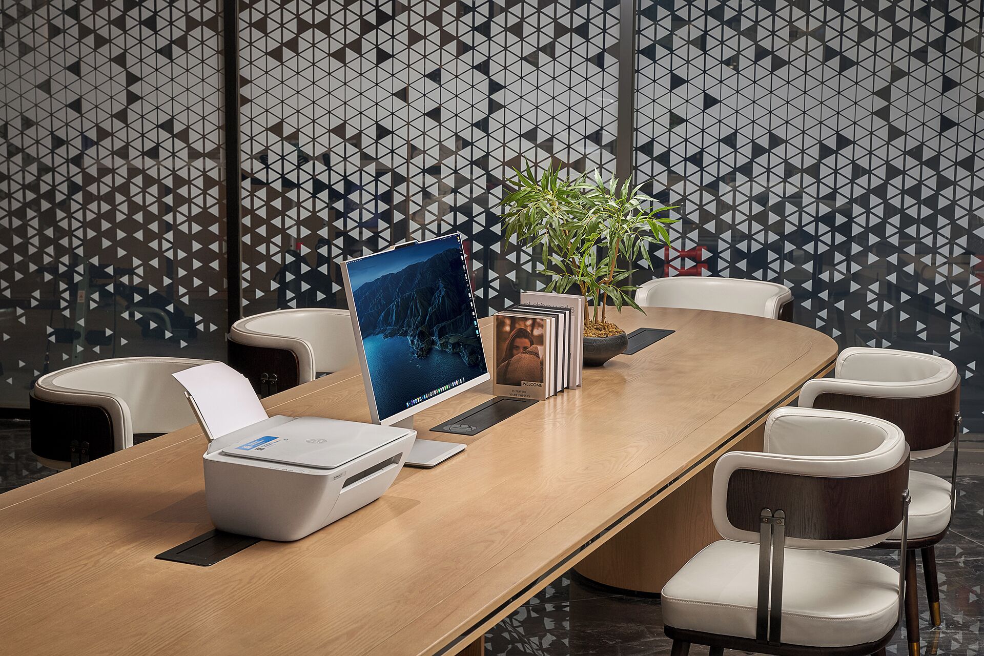 A conference room with a long wooden table, white chairs, a printer, a laptop, a welcome card stand, and a potted plant.