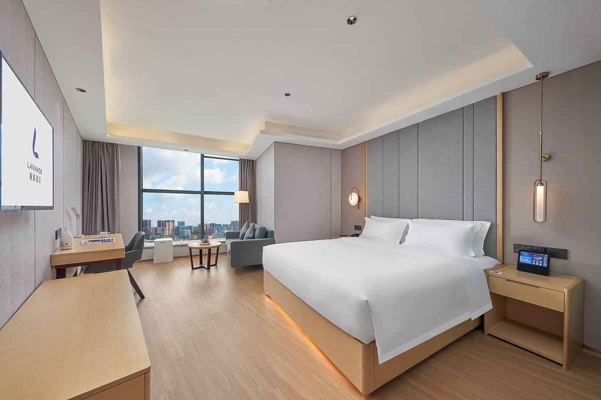 A modern hotel room with a large bed, a desk, a chair, a small table, and a large window offering a city view.