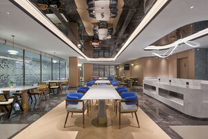 A modern hotel lobby with a long reception desk, multiple seating areas with blue and wooden chairs, and a high ceiling with unique lighting fixtures.