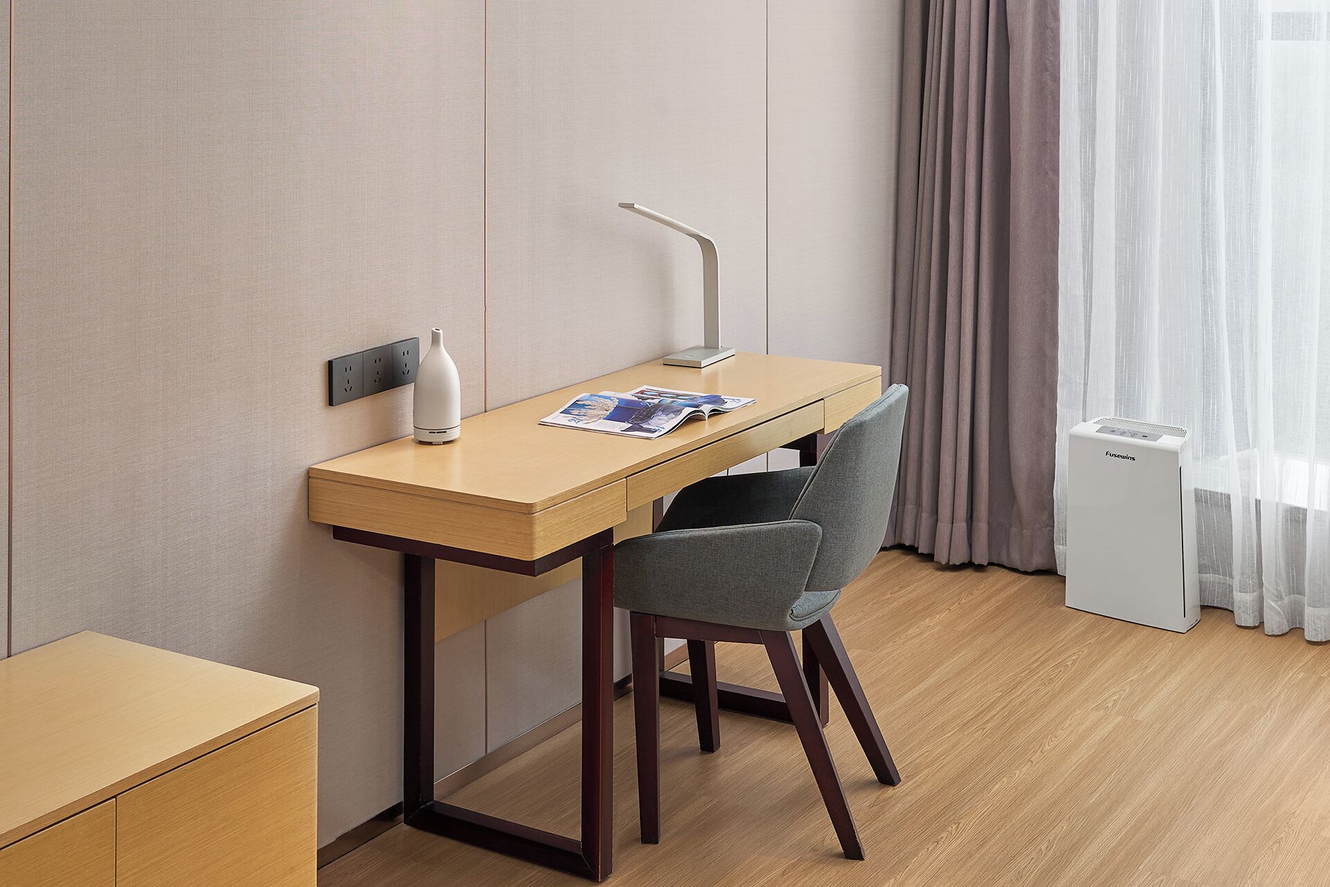A hotel room with a wooden desk, a chair, a small table, a white air purifier, and a window with sheer curtains.