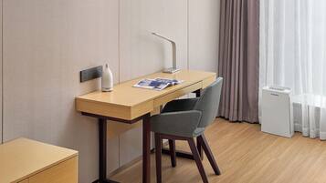 A hotel room with a wooden desk, a chair, a small table, a white air purifier, and a window with sheer curtains.