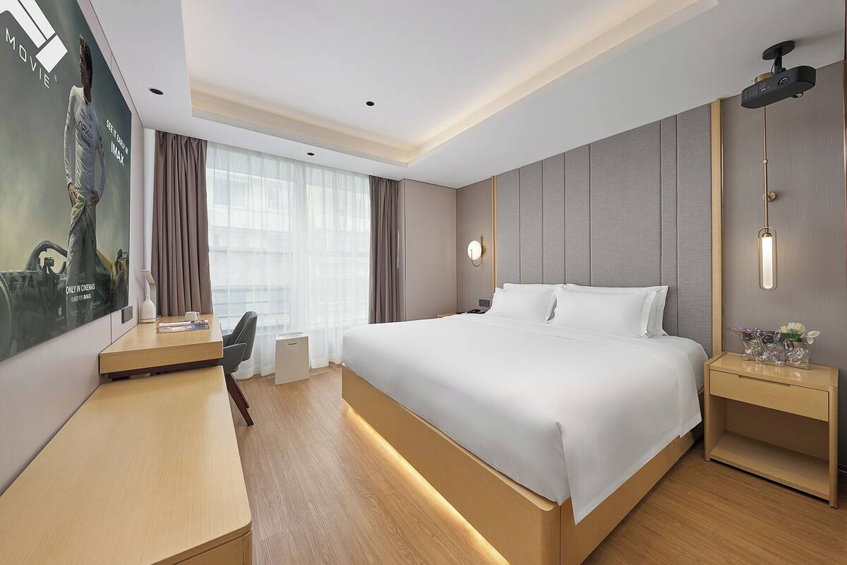 A modern hotel room with a large bed, a desk, a chair, and a TV screen.