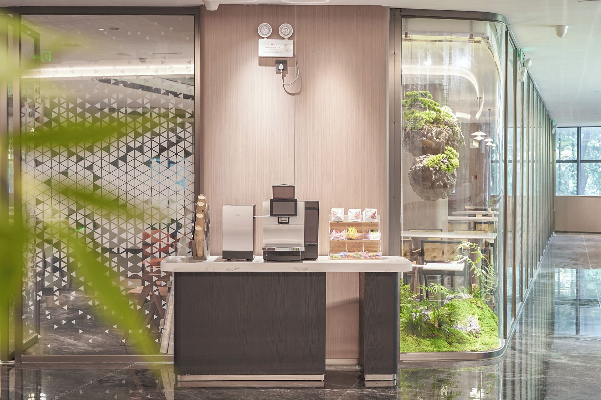 A modern reception desk with a coffee machine, a digital display, and a small plant.