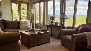 Un salon chaleureux avec un canapé en cuir marron, une table basse en bois et une vue sur la verdure grâce à de grandes fenêtres.