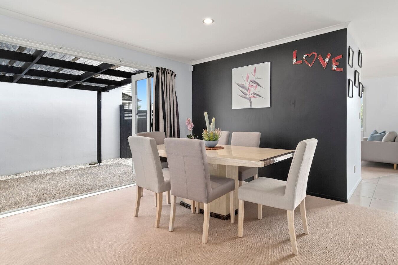 A modern dining area with a wooden table and six chairs, a black accent wall with a 'LOVE' sign, and a view of the outdoors through a glass door.