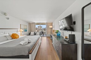 A modern hotel room with a large bed, a flat-screen TV, a desk with a computer, and a view of the city through large windows.