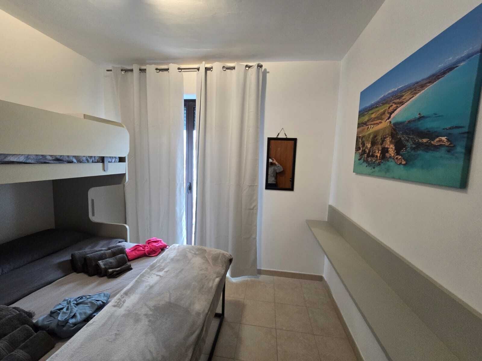 Une pièce comprenant un lit superposé, un canapé, une petite table, un tableau représentant un paysage côtier et une fenêtre avec des rideaux.